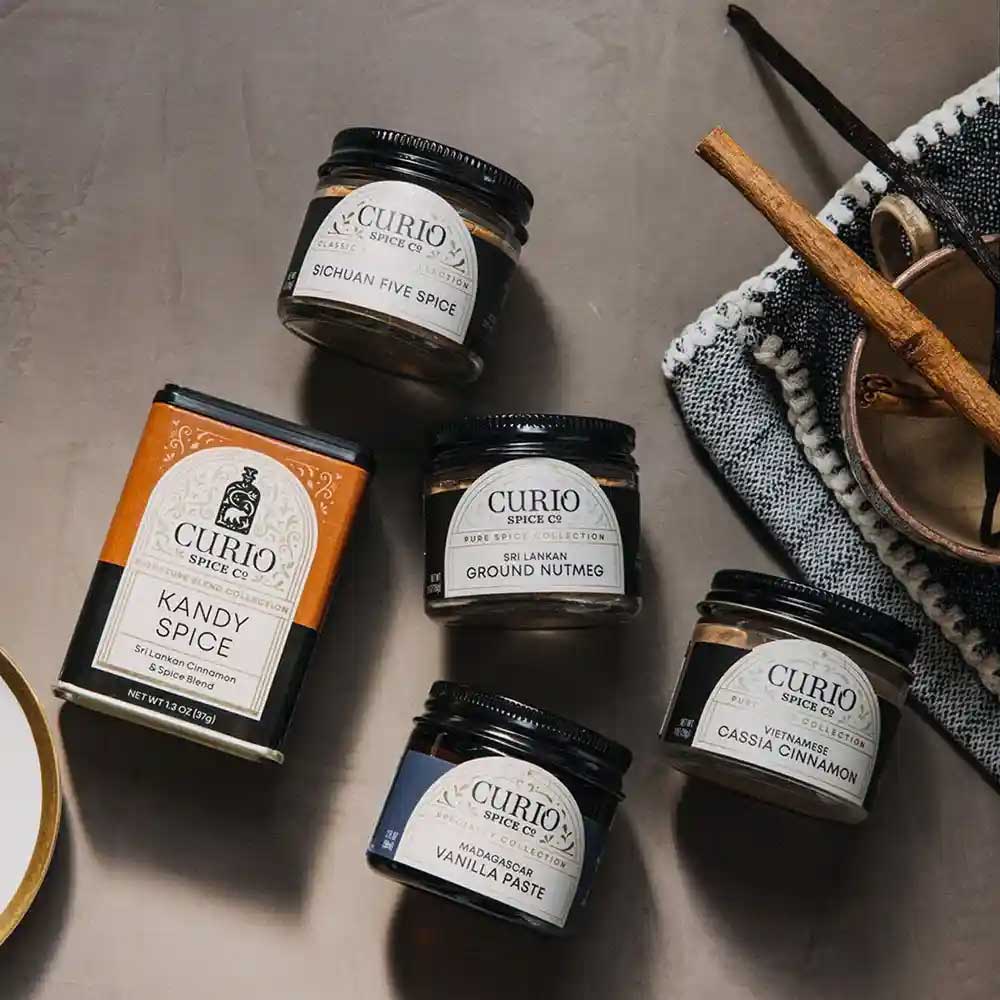 Collection of Curio Spice Co. spice jars on a textured surface with cinnamon sticks.