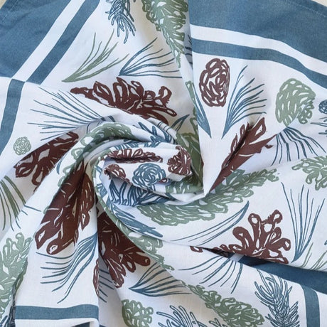 Close-up of bandana with a pinecone and pine needle pattern in brown, green, and blue on a white background.