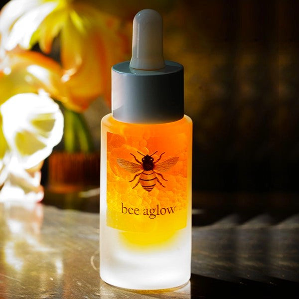 Bee Aglow Facial Serum - a frosted glass bottle filled with a glowing amber serum and bottle dropper top. the bottle has a bee illustration in the center above the text "Bee Aglow"