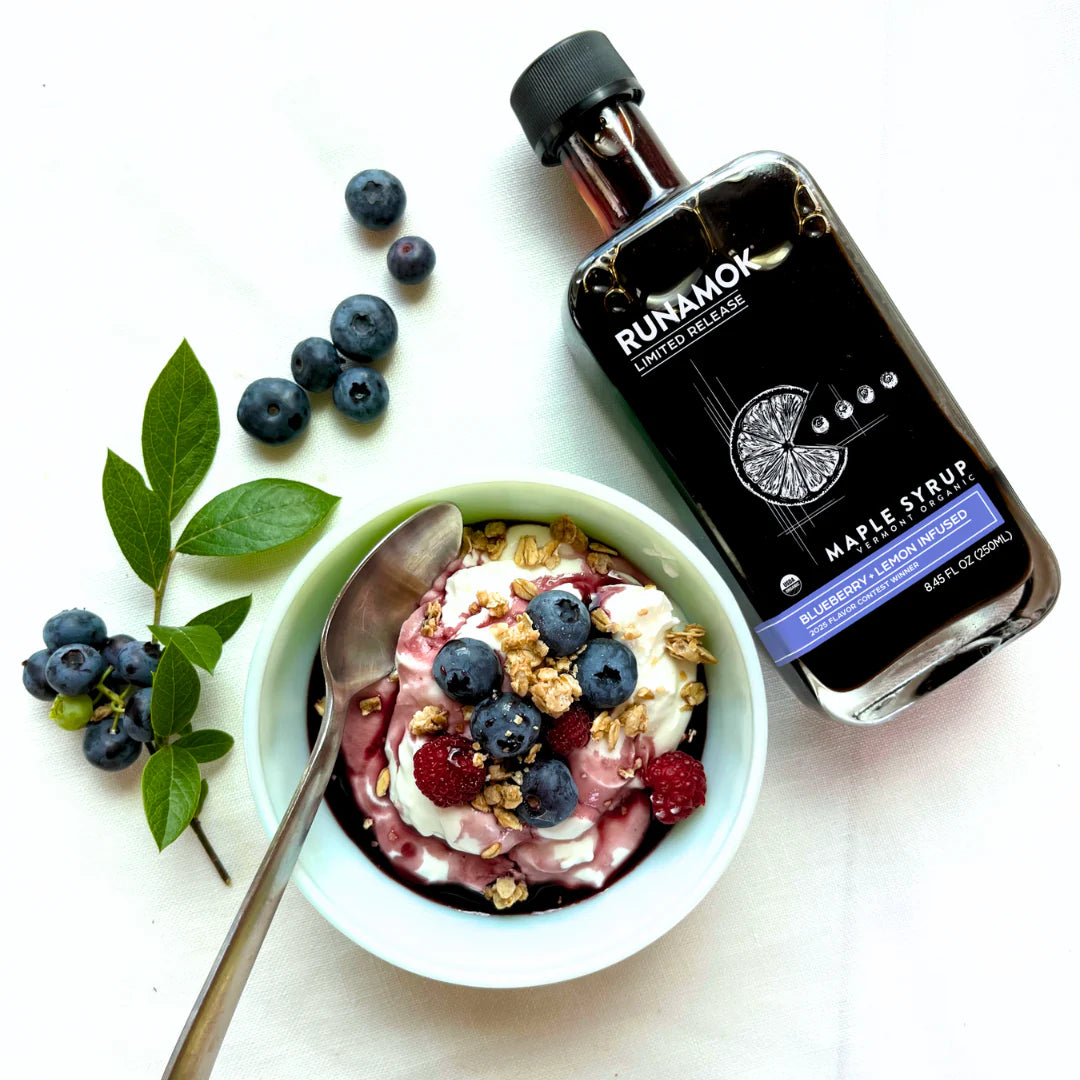 Bowl of yogurt with berries, a spoon, and a bottle of Runamok maple syrup on a white background