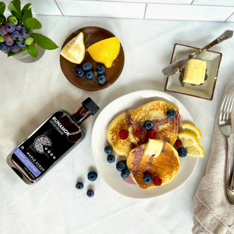 Breakfast setting with pancakes, berries, and a bottle of Runamok Maple syrup on a white surface.