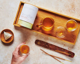 Tea-making setup with a wooden tray, tea bags, and a bottle labeled 'Breathe' on a textured surface.