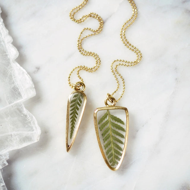 a necklace featuring a preserved fern pressed inside clear resin, framed by a delicate 24kt gold-plated arrow-shaped pendant with a gold bead chain laid on a white marble surface