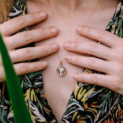 a close up of a model wearing the Pressed Fern Arrow Necklace with the models hands gently touching the edge of the gold chain showing that the pendant is about the length of her fingernail
