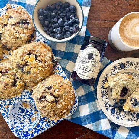a table scene with a jar of blueberry jam and a bowl of blueberries alongside a pan of blueberry muffins next to a plate with a sliced blueberry muffin
