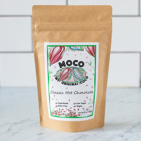 a kraft hot cocoa package with a white label with red and green illustrations of cocoa beans that reads "MOCO My Original Coco" on a marble counter with white subway tile background
