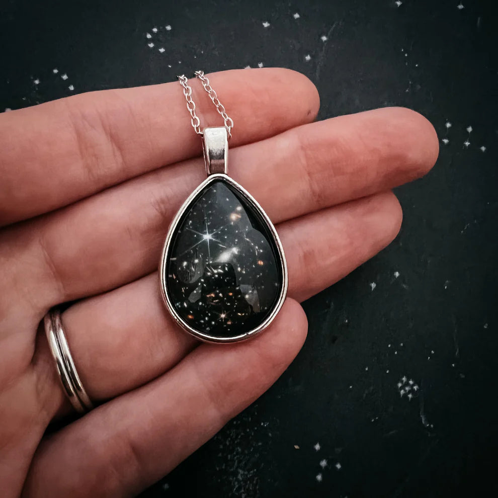 Teardrop-shaped pendant with celestial design held in a hand against a dark background