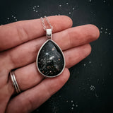 Teardrop-shaped pendant with celestial design held in a hand against a dark background