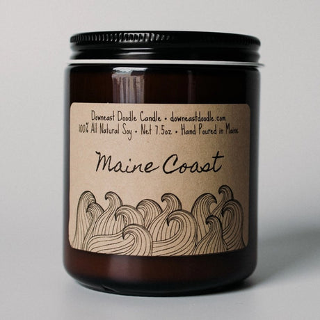 a candle in an amber glass jar with black lid and kraft label with handwriting style graphics that read "Maine Coast" above playful doodle style line drawings of ocean waves