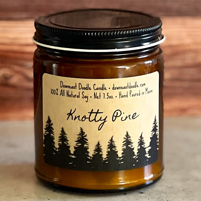 a candle in an amber glass jar with black lid and kraft label with handwriting style graphics that read "Knotty Pine" above a silhouetted forest of pine trees