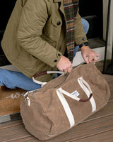 a model kneeling near a doorway unzipping their brown waxed canvas duffle bag