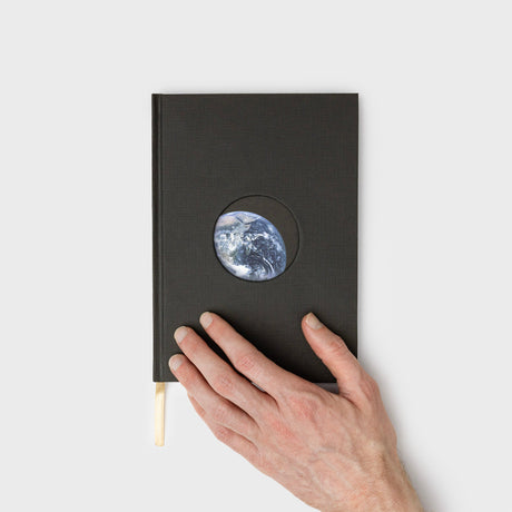 Black notebook with a circular cutout showing Earth, held by a hand on a light gray background