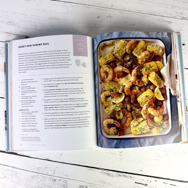 Open cookbook with a sheet pan of shrimp boil on a wooden surface