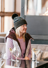 A woman wearing a dark knit beanie with a pom-pom and a brown and white jacket sits at a table in a restaurant or bar, with cocktail glasses in front of her.