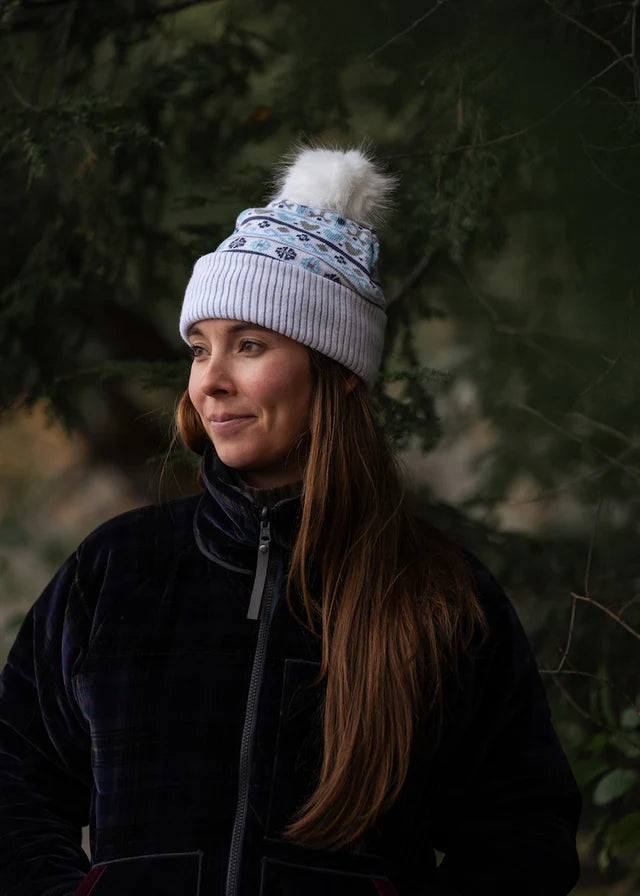 A woman wearing a light gray ribbed knit beanie with a white faux fur pom-pom and a blue patterned band, paired with a dark navy fleece jacket, set against a dark, blurred forest background.