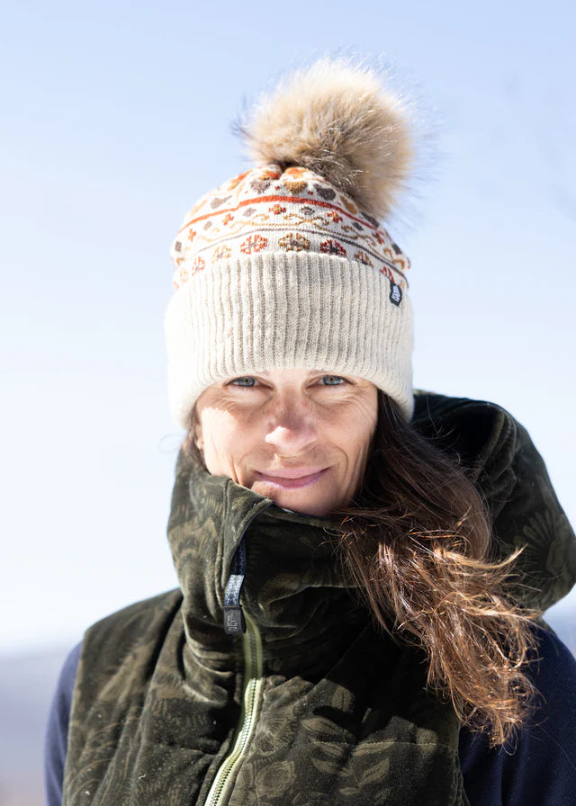 A woman outdoors wearing a cream knit beanie with a brown faux-fur pom-pom and a patterned band, along with a dark olive green insulated jacket against a pale blue sky.