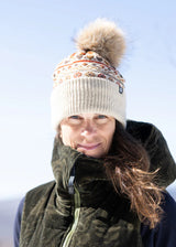 A woman outdoors wearing a cream knit beanie with a brown faux-fur pom-pom and a patterned band, along with a dark olive green insulated jacket against a pale blue sky.