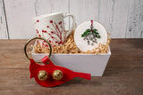 the festive accents gift box on a wood table - the winterberries mug and mistletoe ornament are arranged in a white tray surrounded by kraft colored crinkle packing and the cardinal door hanger bell sits in front