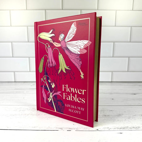 Book titled 'Flower Fables' by Louisa May Alcott with a fairy illustration on a magenta cover with gold edged pages against a white tiled wall.