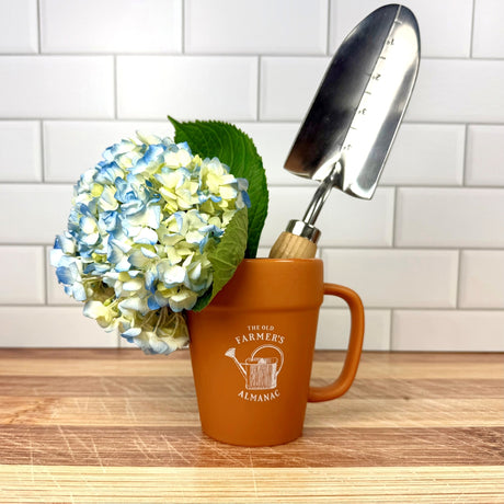 terracotta style mug with 'The Old Farmer's Almanac' logo and vintage watering can graphic with hydrangea blossoms and a garden trowel in it
