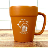 terracotta style mug with 'The Old Farmer's Almanac' logo and vintage watering can graphic on a wooden surface.