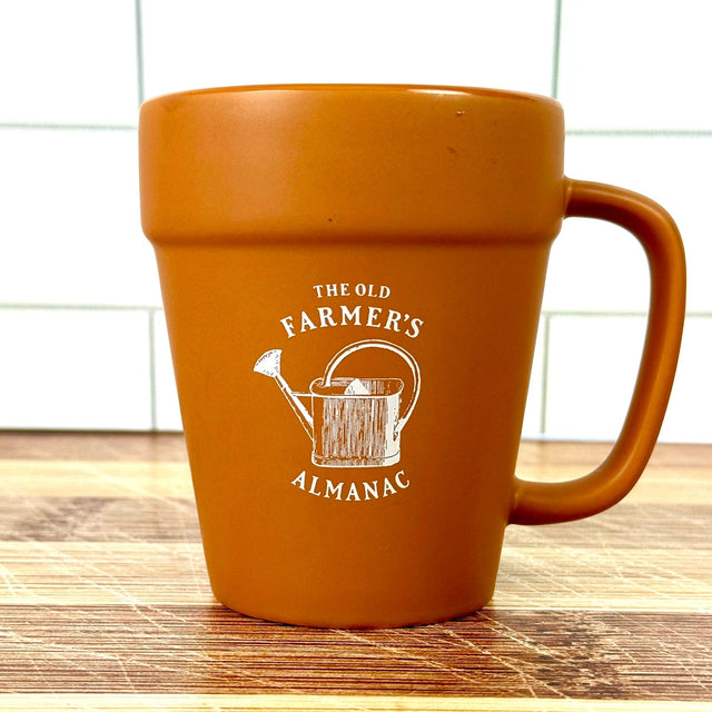 terracotta style mug with 'The Old Farmer's Almanac' logo and vintage watering can graphic on a wooden surface.