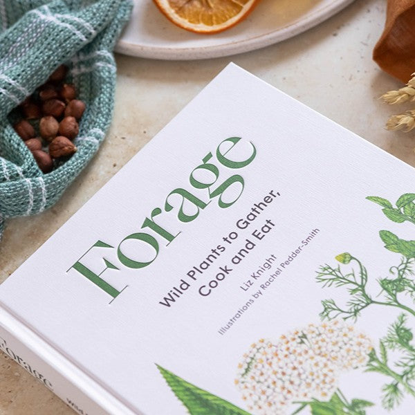 Book titled 'Forage: Wild Plants to Gather, Cook and Eat' on a surface with herbs and an orange slice.