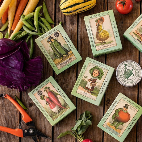 Vintage-style gardening soap packaging with illustrations of turn of the century ladies and gentlemen dressed as various vegetables on a wooden surface with vegetables and tools.