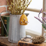 A decorative beehive-shaped wall hanging with bees and honeycomb details, displayed in front of a corrugated metal bucket filled with willow branches on a rustic wooden surface.