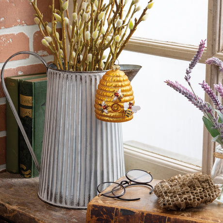 A decorative beehive-shaped wall hanging with bees and honeycomb details, displayed in front of a corrugated metal bucket filled with willow branches on a rustic wooden surface.