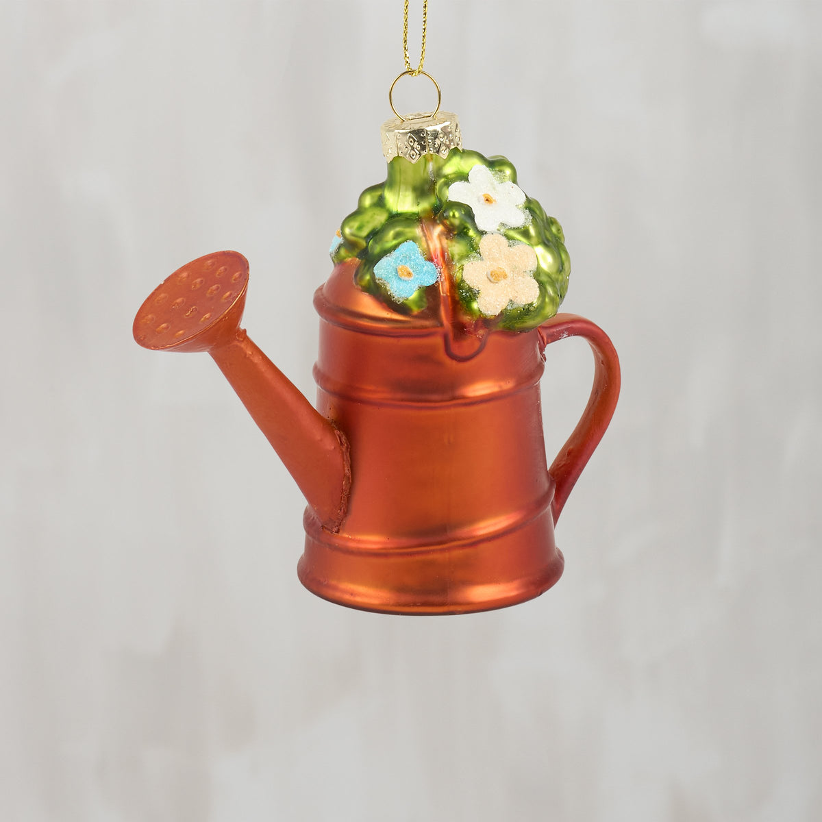 A Christmas ornament shaped like a metallic copper watering can with a green top and small white and blue flowers, hanging against a light gray background.