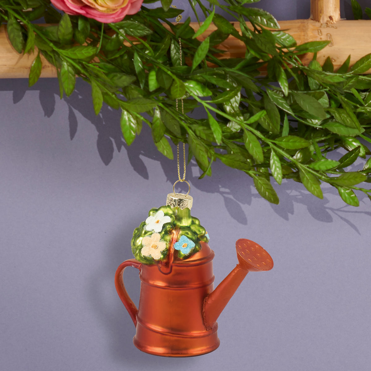 A decorative Christmas ornament shaped like a copper-colored watering can with a green top featuring white and yellow flowers, hanging from a gold string beneath artificial green leaves.