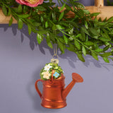 A decorative Christmas ornament shaped like a copper-colored watering can with a green top featuring white and yellow flowers, hanging from a gold string beneath artificial green leaves.