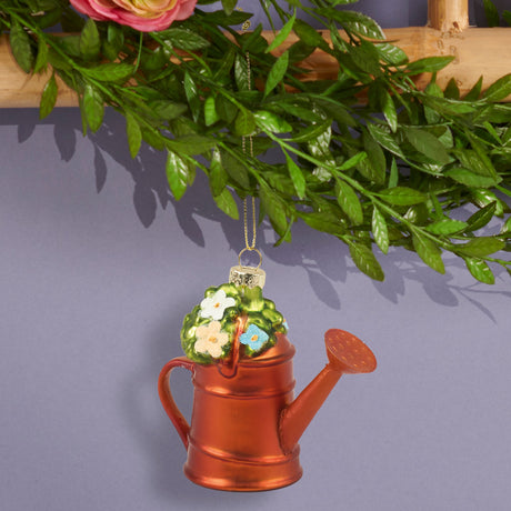 A decorative Christmas ornament shaped like a copper-colored watering can with a green top featuring white and yellow flowers, hanging from a gold string beneath artificial green leaves.