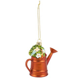 A copper-colored watering can Christmas ornament decorated with green leaves and white and light blue flowers, hanging from a gold string on a white background.