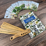 The Old Farmer's Almanac Guide to Herbs on a wood surface alongside Old Farmer's Almanac Plant Markers, a spread of six herb seed packets, and a wooden herb stripper