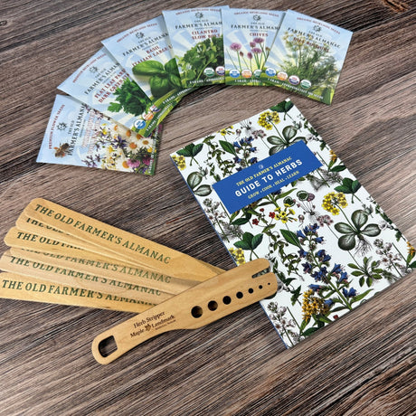 The Old Farmer's Almanac Guide to Herbs on a wood surface alongside Old Farmer's Almanac Plant Markers, a spread of six herb seed packets, and a wooden herb stripper