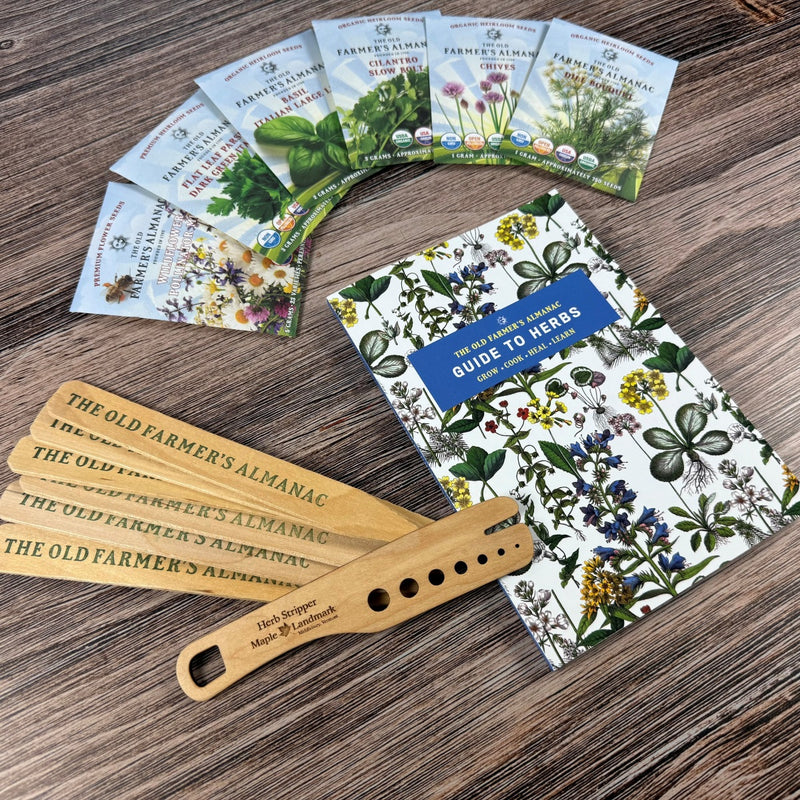 The Old Farmer's Almanac Guide to Herbs on a wood surface alongside Old Farmer's Almanac Plant Markers, a spread of six herb seed packets, and a wooden herb stripper