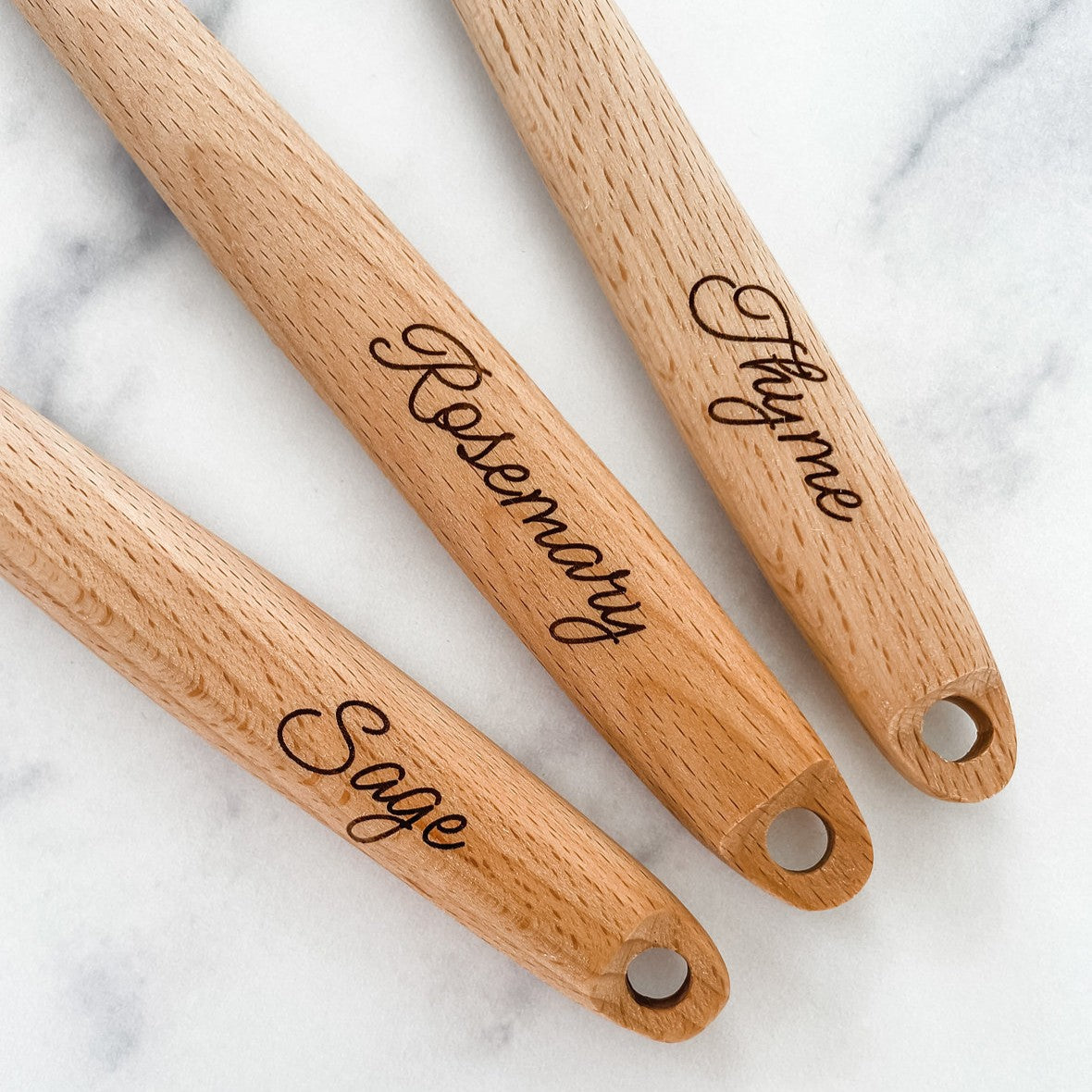 the handles of three wooden kitchen utensils with engraved herb names on a marble surface - Sage, Rosemary, and Thyme
