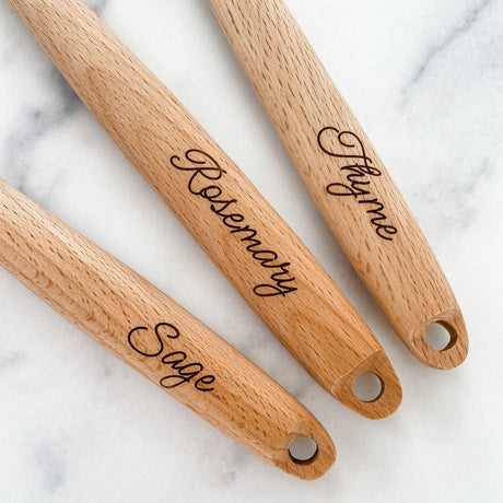 the handles of three wooden kitchen utensils with engraved herb names on a marble surface - Sage, Rosemary, and Thyme