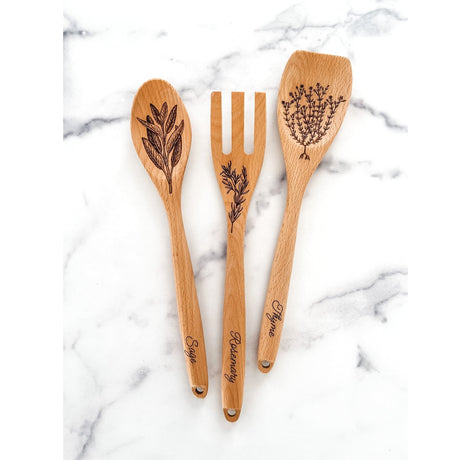 Three wooden utensils with engraved herb designs on a marble surface - featuring Sage, Rosemary, and Thyme