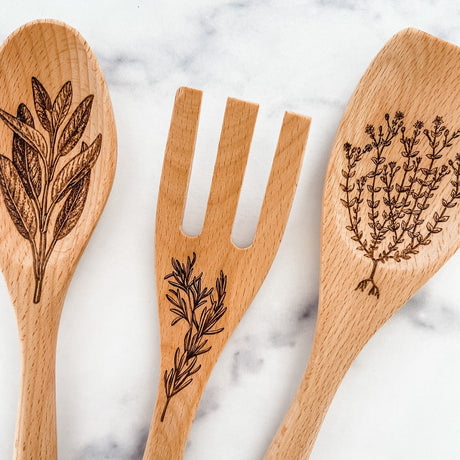 Close up of three wooden utensils with engraved herb designs on a marble surface