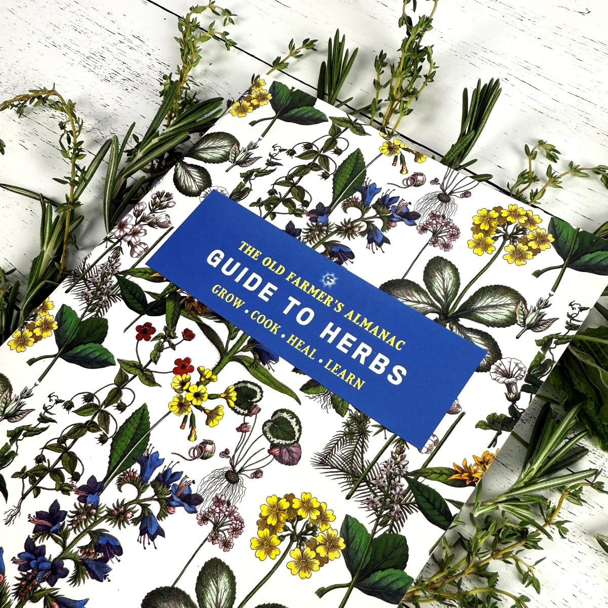 Book titled 'The Old Farmer's Almanac Guide to Herbs' surrounded by herbs and flowers on a white background