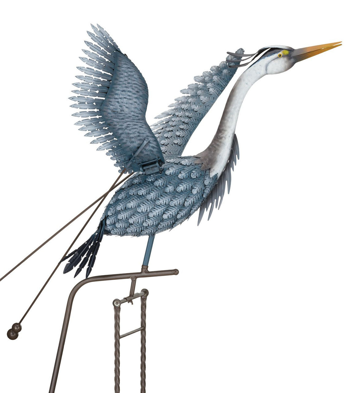 Metal sculpture of a heron in mid-flight, with wings extended upward and long neck outstretched. The sculpture is designed with intricate, feather-like details and is mounted on a metal rod with counterweights. The bird's body is painted in blue and gray tones, and its long beak points forward.