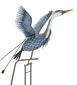 Metal sculpture of a heron in mid-flight, with wings extended upward and long neck outstretched. The sculpture is designed with intricate, feather-like details and is mounted on a metal rod with counterweights. The bird's body is painted in blue and gray tones, and its long beak points forward.