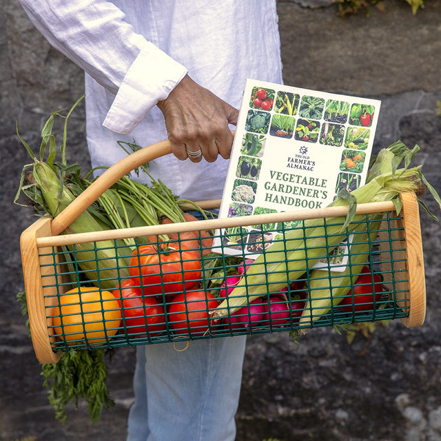 Person holding a wood and wire garden hod full of fresh vegetables with a 'Vegetable Gardener's Handbook' inside.