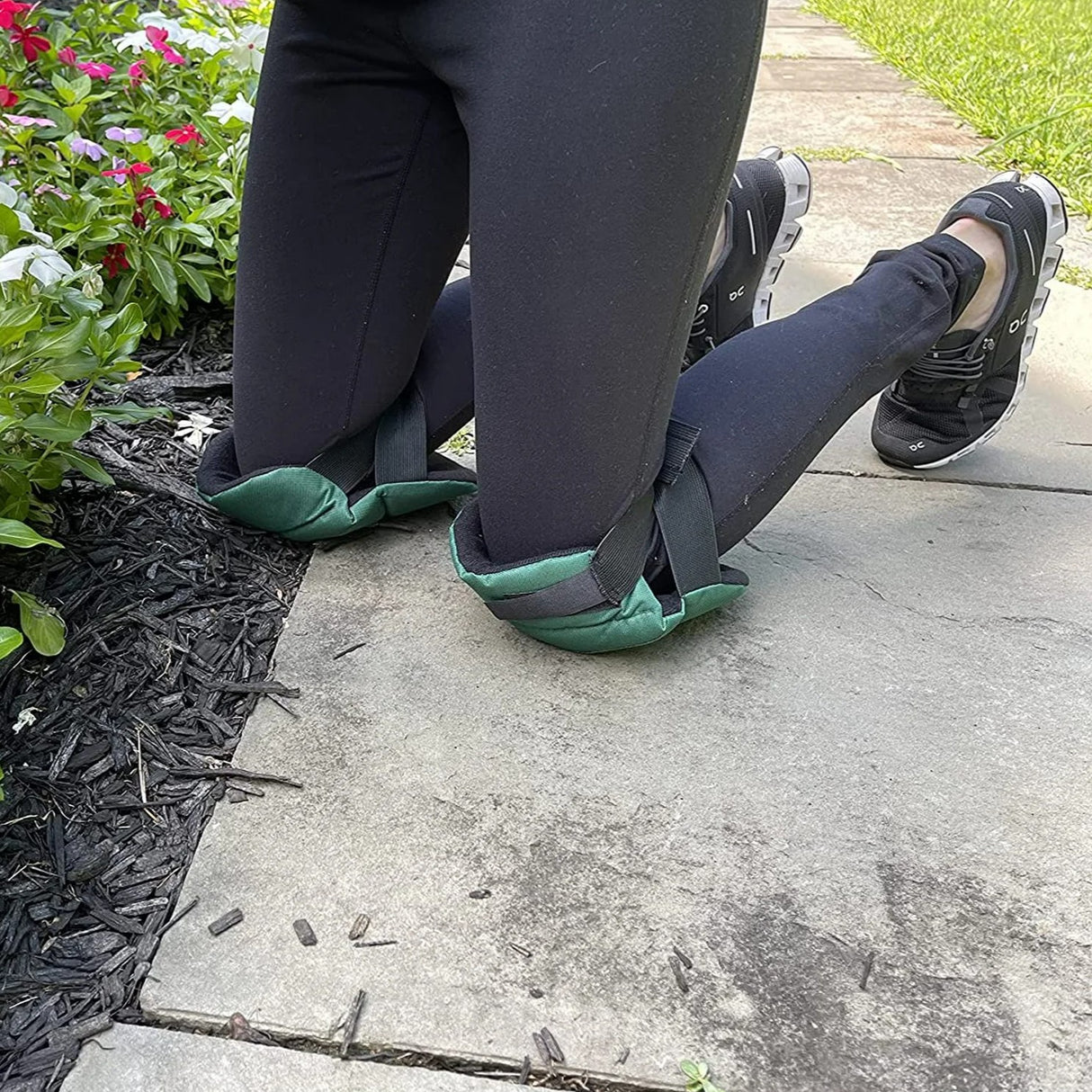 Person wearing green kneepads kneeling on a sidewalk with flowers and grass in the background