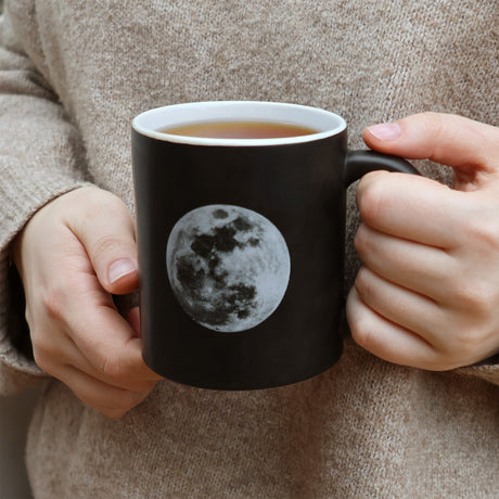 Person holding a black mug with a moon design, filled with a warm beverage.