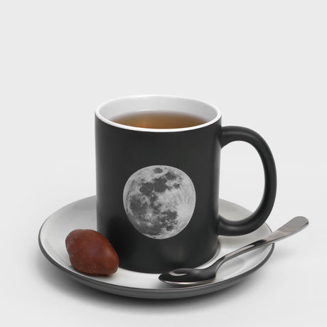 Black mug with moon design on a white saucer with a spoon and treat on a white background