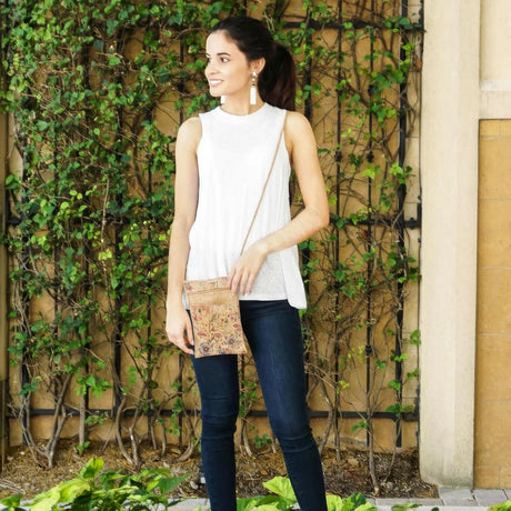 Woman in white sleeveless top and blue jeans holding a wildflower print crossbody bag against a green ivy wall